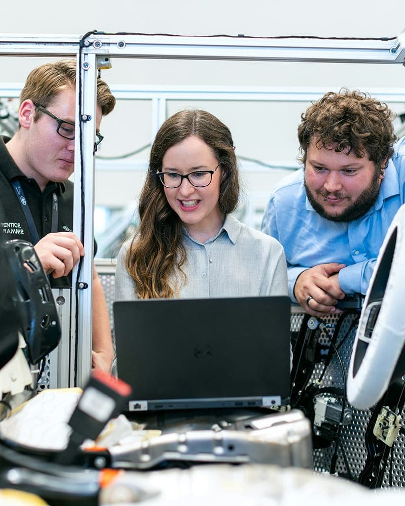 Three colleagues gathered around a laptop beside engineering equipment, reviewing the screen together